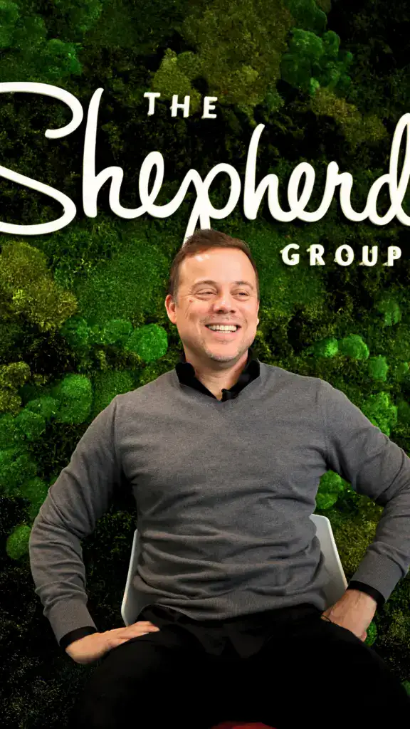 Man smiling in front of a green, leafy background with "The Shepherd Group" text.