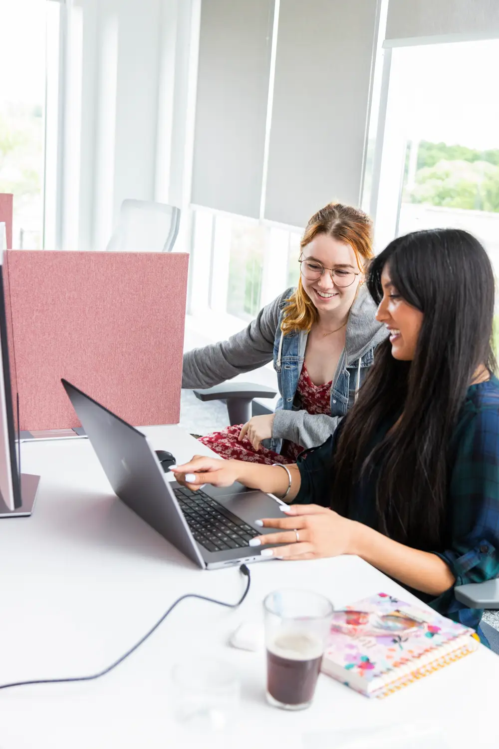 Two women collaborating on a laptop in a bright office setting.