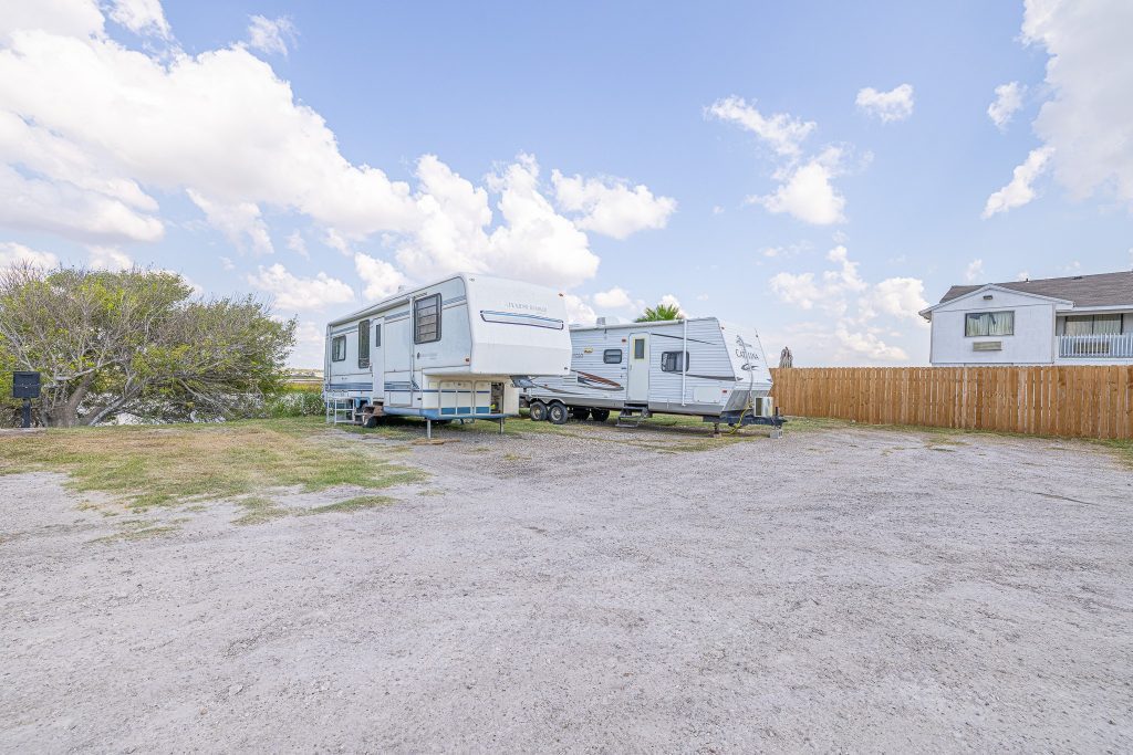 Portland, TX RV Park Two white RVs parked at Portland RV Park in Portland, Texas, on gravel lot near a wooden fence, tree, and house with cloudy sky.