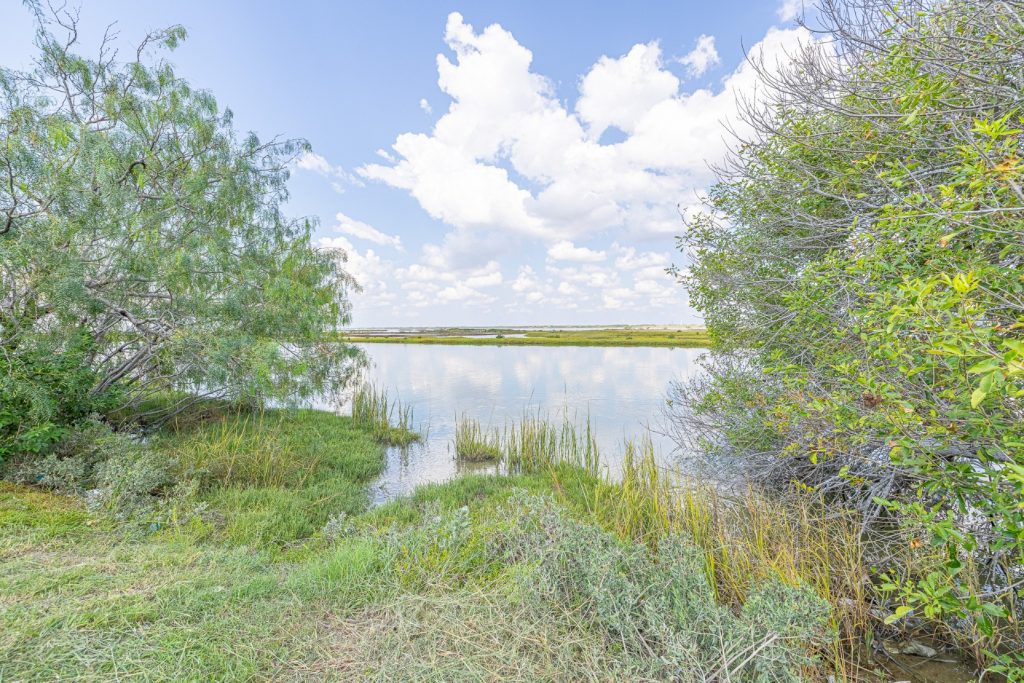 Portland, TX RV Park Serene lakeside view near Portland RV Park in Portland, Texas, with green bushes, calm water, cloudy sky reflection, and distant fields.
