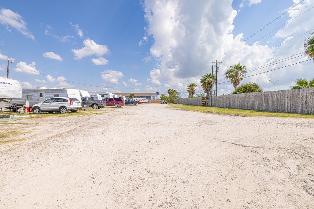 Portland, TX RV Park Spacious sandy parking lot at Portland RV Park in Portland, Texas with cars, RVs, palm trees, and wooden fence under blue sky.