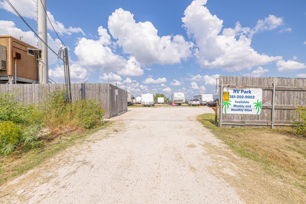 Portland, TX RV Park Gravel driveway entering Portland RV Park in Portland, Texas, with RVs, wooden gate, sign for weekly/monthly sites, blue sky.
