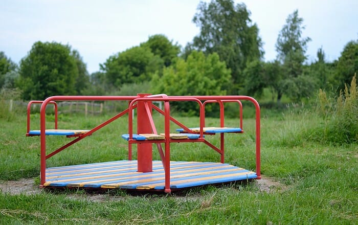 Rotes Karussell auf einem Spielplatz mit grüner Wiese und Bäumen im Hintergrund.