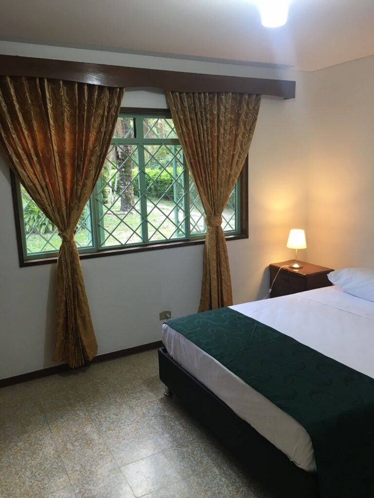 Arreglo de habitación de hotel con cama doble, cortinas de tela dorada, ventana con rejas y vista a un jardín verde, lámpara de mesa encendida, ambiente acogedor y tranquilo.