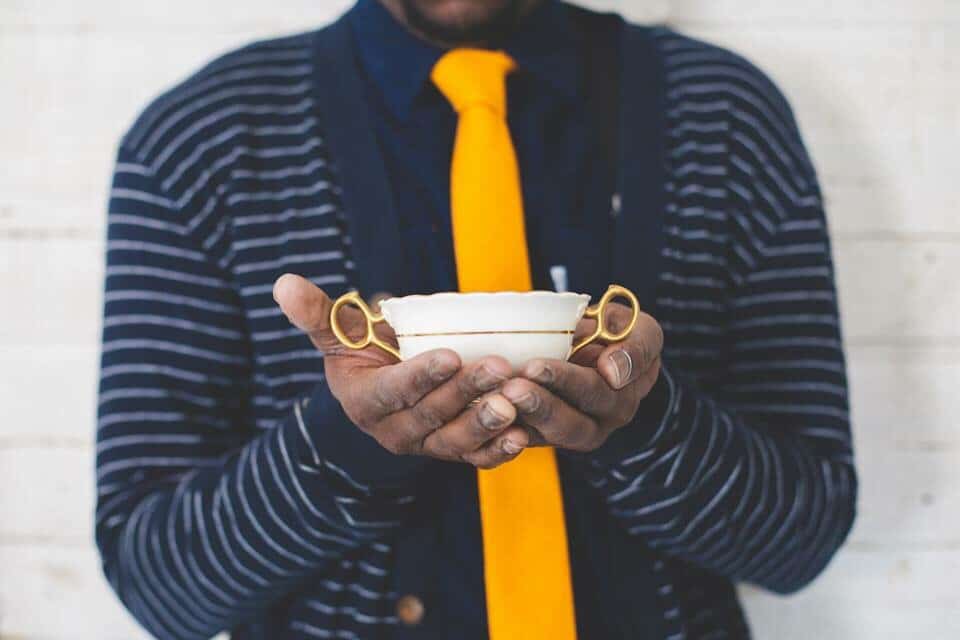 A person in a striped cardigan and yellow tie holds a white and gold teacup with both hands