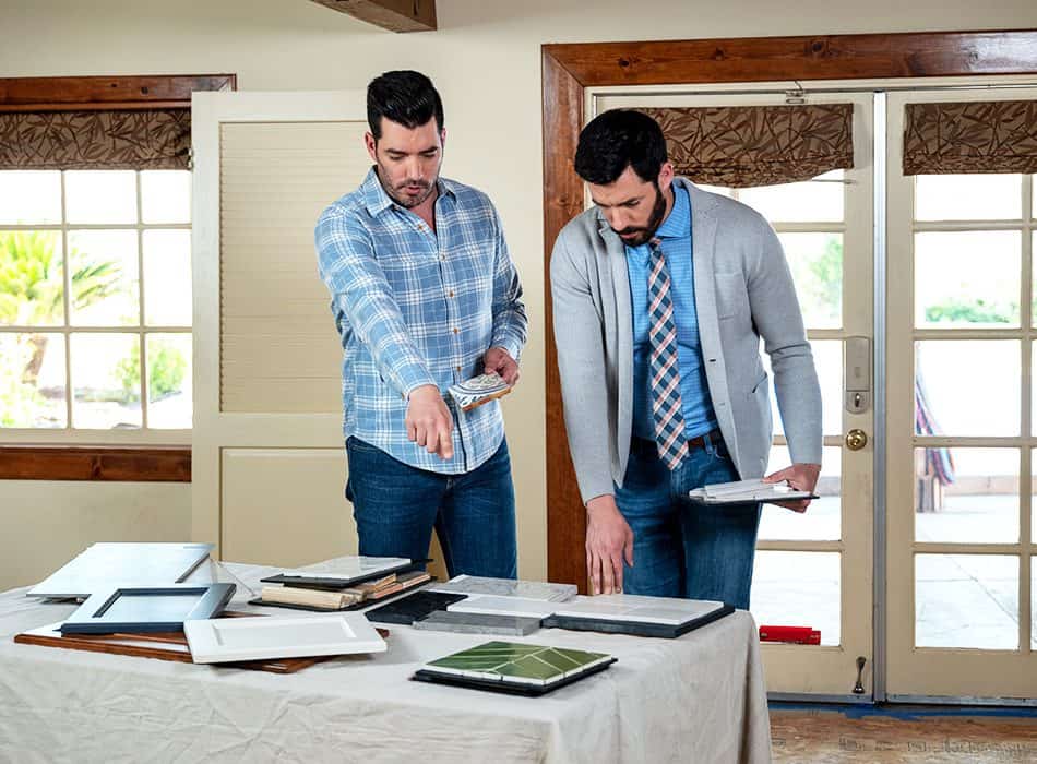 Drew and Jonathan Scott reviewing tile and frame samples on a table in a bright room.