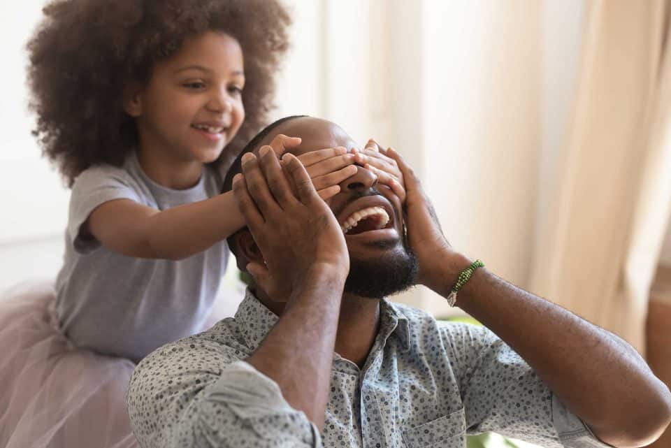 Smiling father with a beard sitting while his daughter playfully covers his eyes from behind