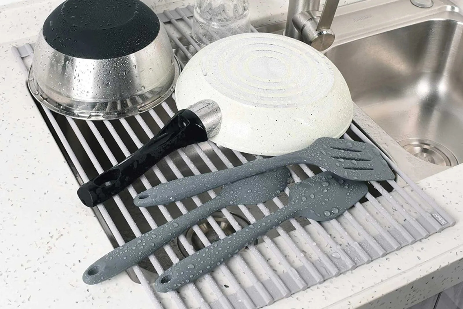 Wet dishes, utensils, and a frying pan drying on a roll-up dish rack over a kitchen sink