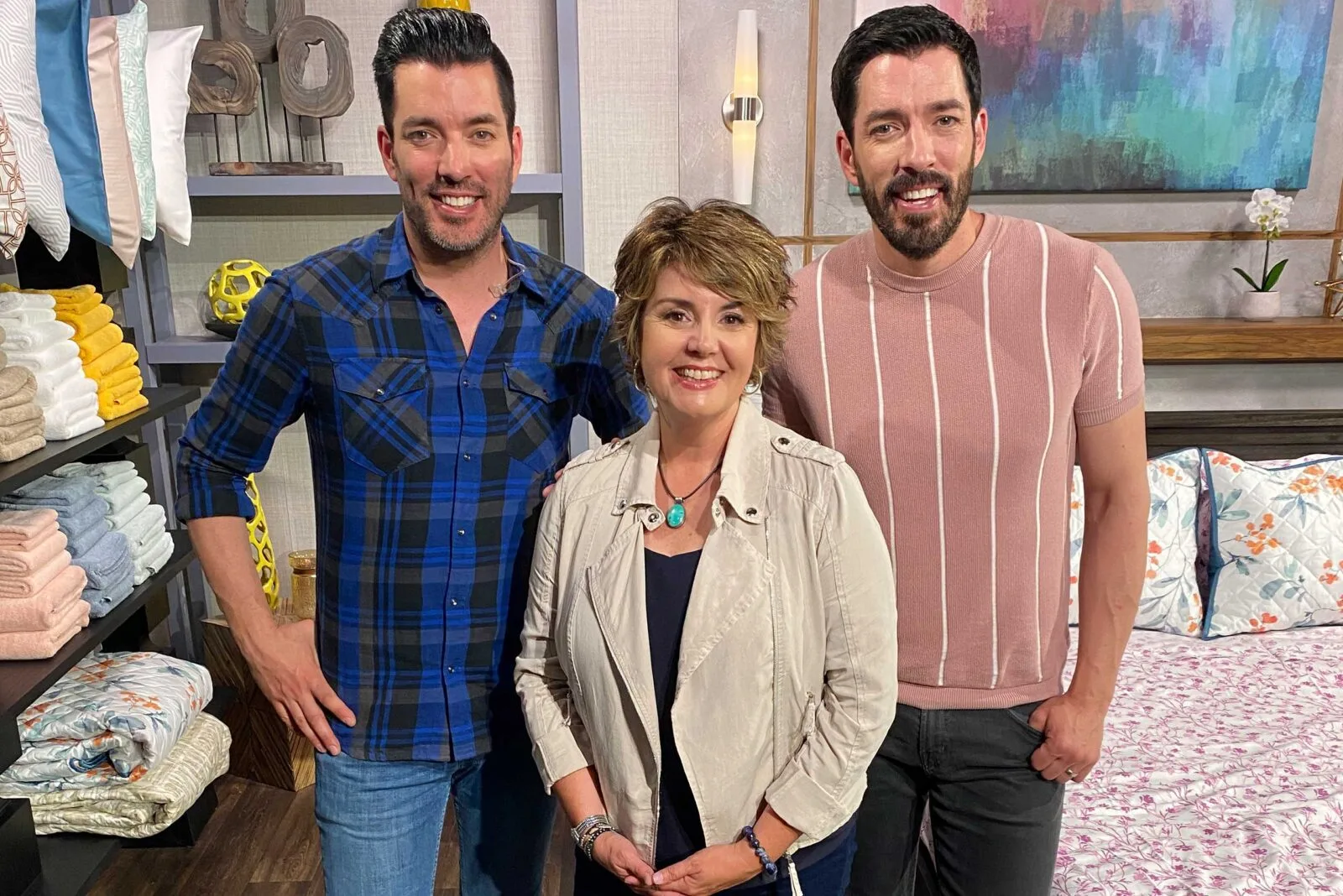 Scott brothers and a woman posing together in a home decor setting with folded towels and bedding in the background