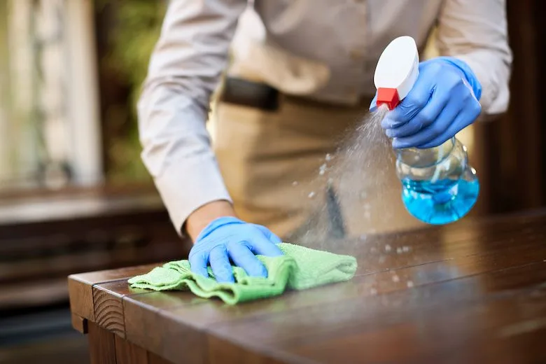 Person cleaning a wooden table with a green cloth and blue spray cleaner while wearing blue gloves