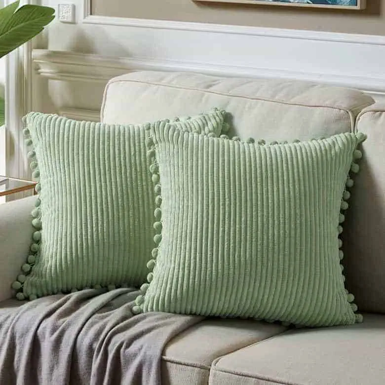 Two green ribbed throw pillows with pom-pom trim on a beige sofa