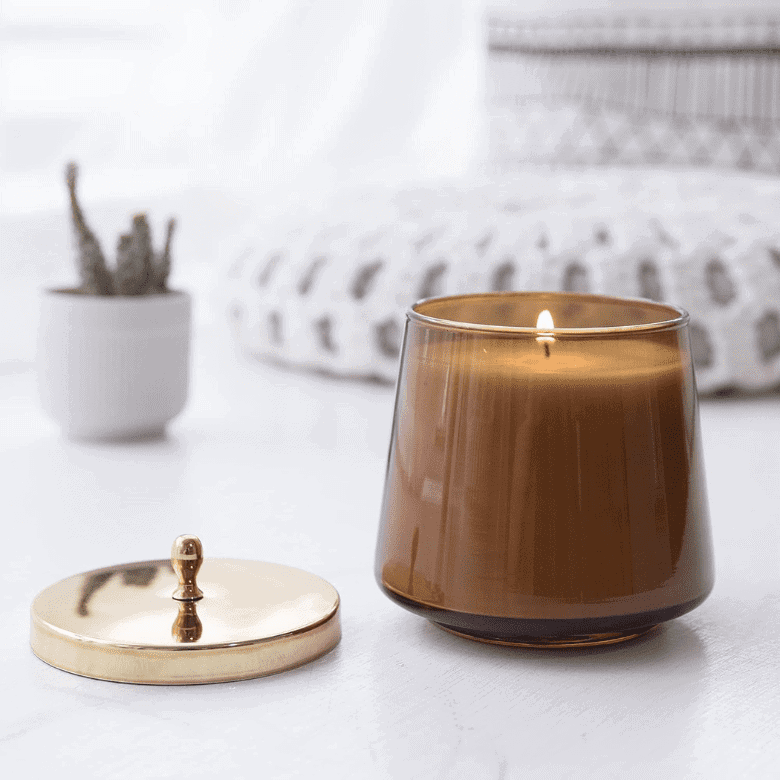 Amber glass candle with gold lid on a white surface, soft decor background