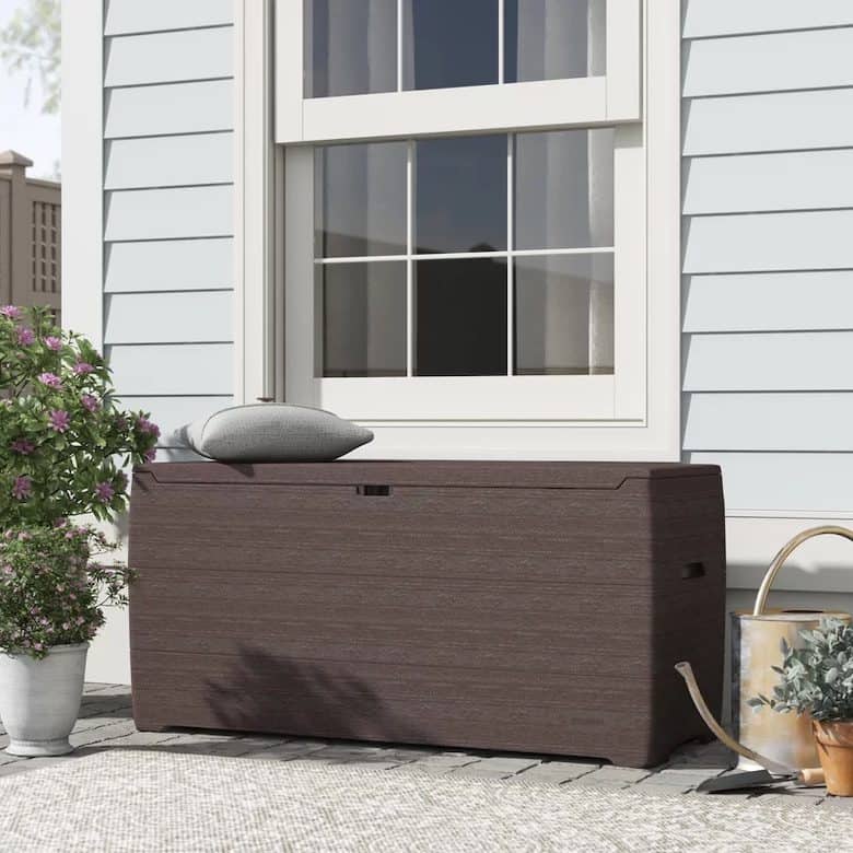 Outdoor storage bench with a pillow, next to a potted plant and watering can, in front of a window