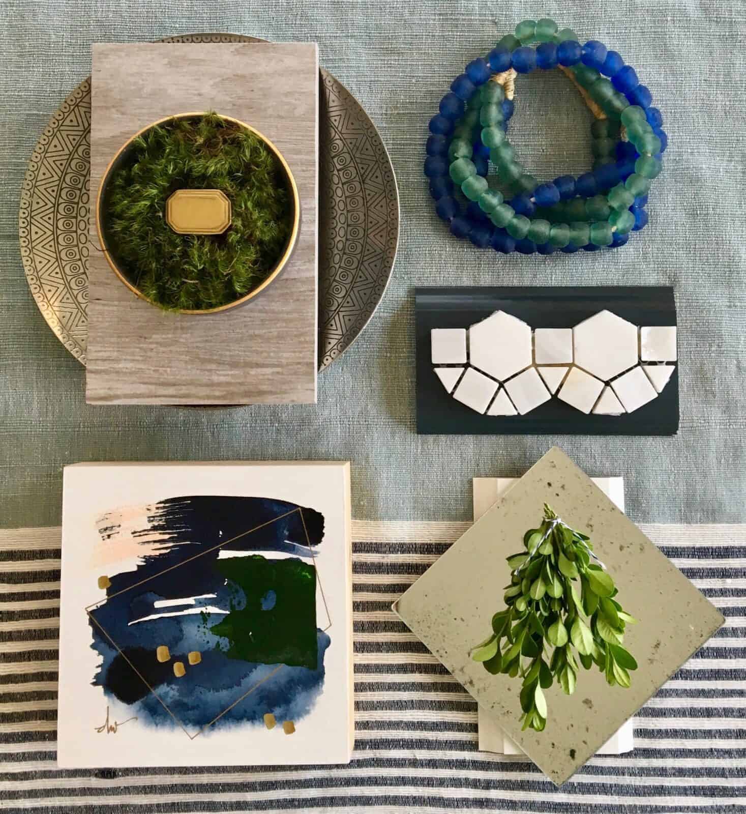 Decorative flat lay with textured tiles, green foliage, blue beads, and abstract art pieces