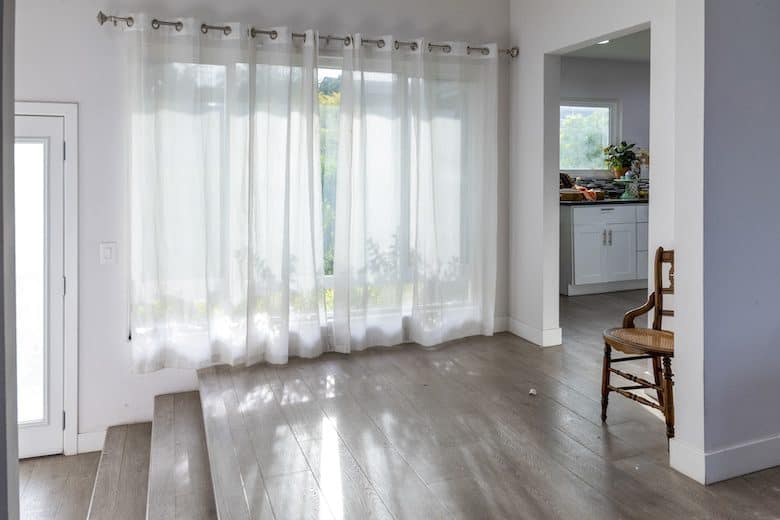 Bright room with sheer white curtains, wooden flooring, and a simple wooden chair by the doorway