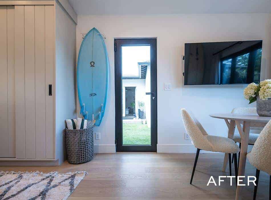 Modern coastal living space with a mounted surfboard, black-framed glass door, wall-mounted TV, and dining area