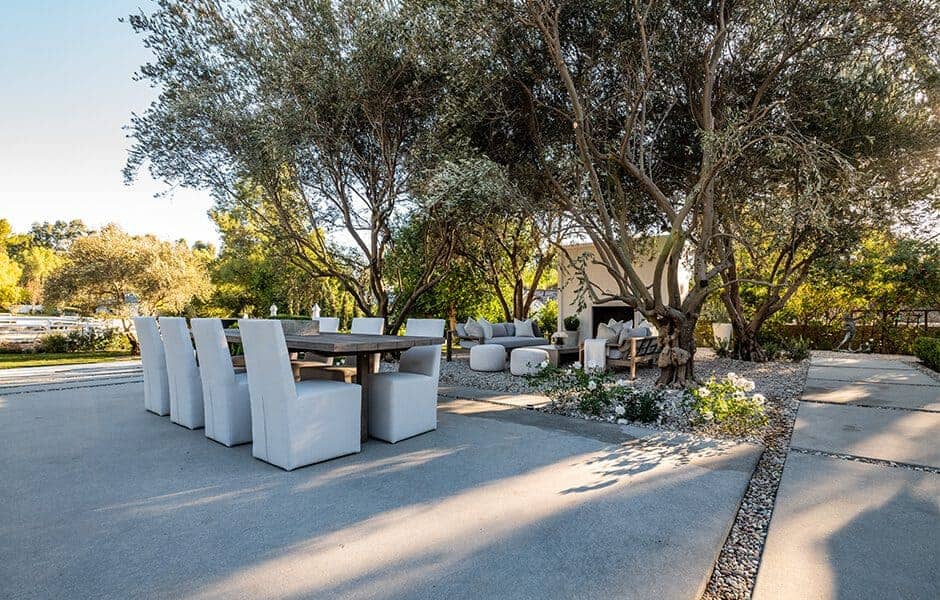 Outdoor dining and lounge area surrounded by trees and greenery on a concrete patio