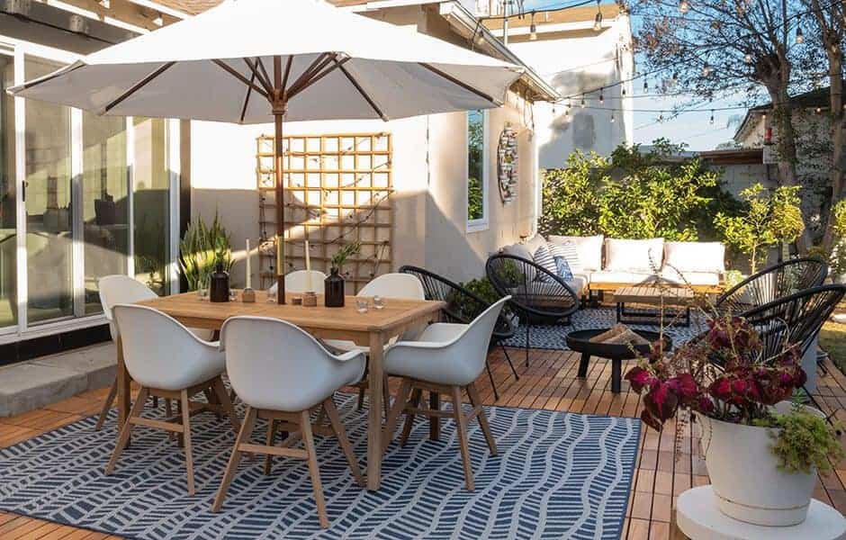 Outdoor patio with a wooden dining table, white chairs, large umbrella, seating area, and vibrant plants on a patterned rug