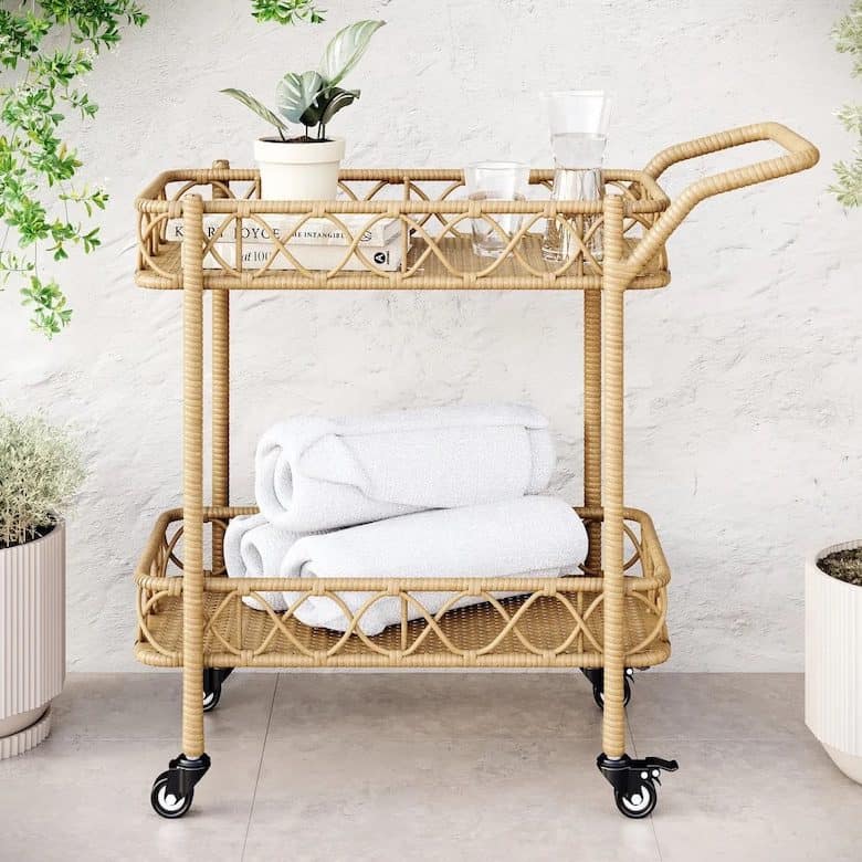 Rattan bar cart with wheels, holding glassware, books, a potted plant, and folded white towels