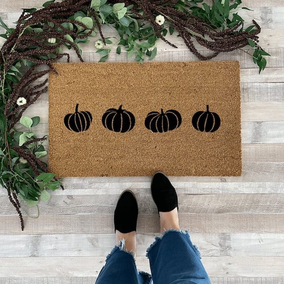 A coir doormat with four pumpkin silhouettes, surrounded by greenery