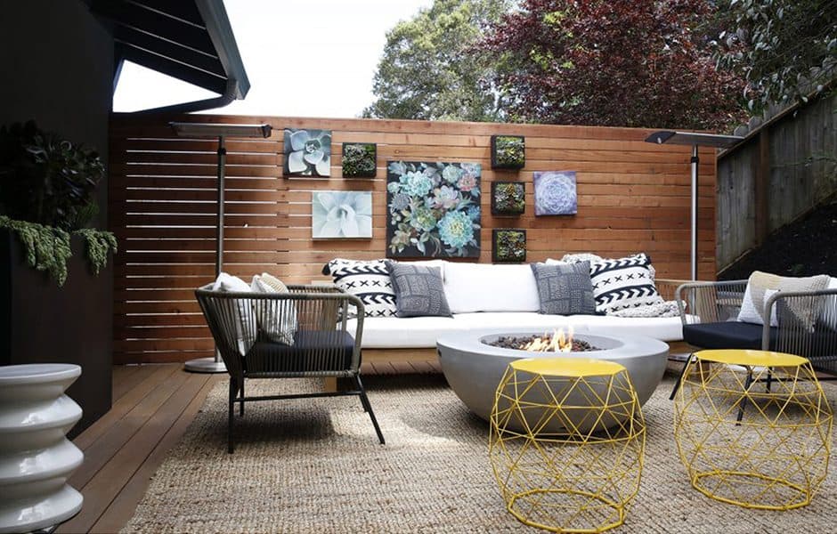 Outdoor seating area with sectional couch, fire pit, yellow side tables, and floral wall art