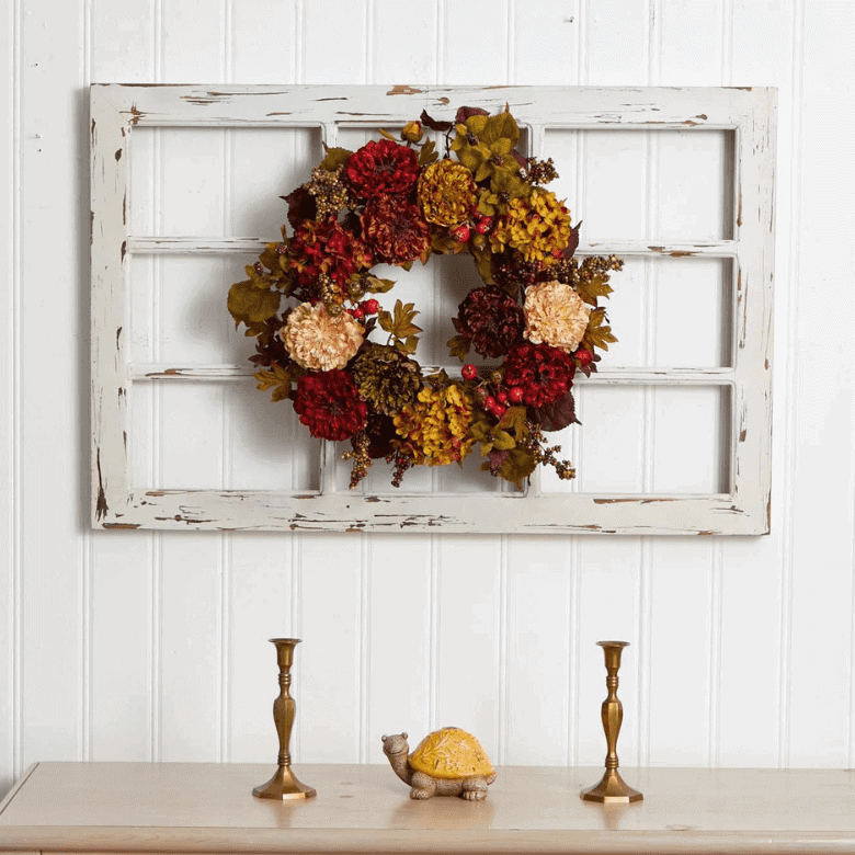 Decorative fall wreath with red, yellow, and orange flowers hanging on a distressed white window frame