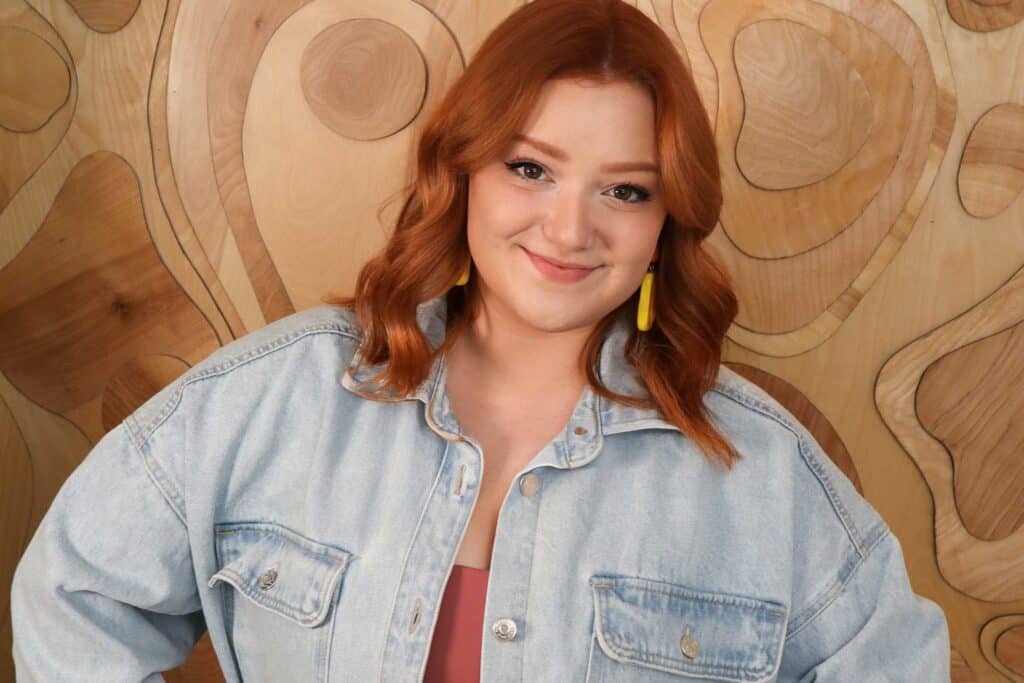 Smiling woman with red hair wearing a denim jacket and yellow earrings against a wood-patterned background