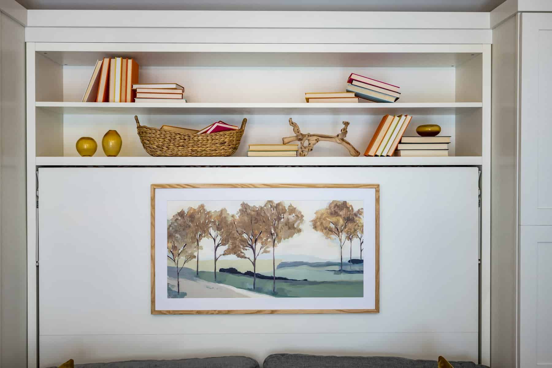 White built-in bookshelf with books, decorative items, and a framed landscape painting on a fold-down bed