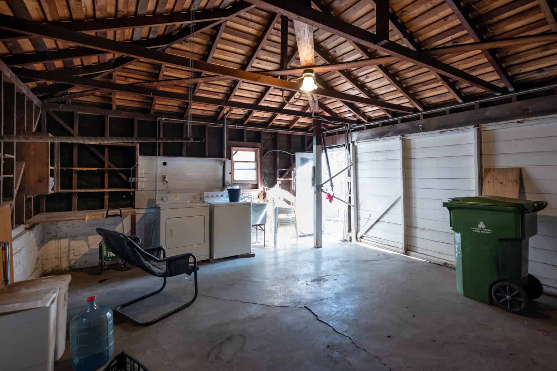 Dimly lit garage with exposed beams, washer-dryer unit, and scattered storage