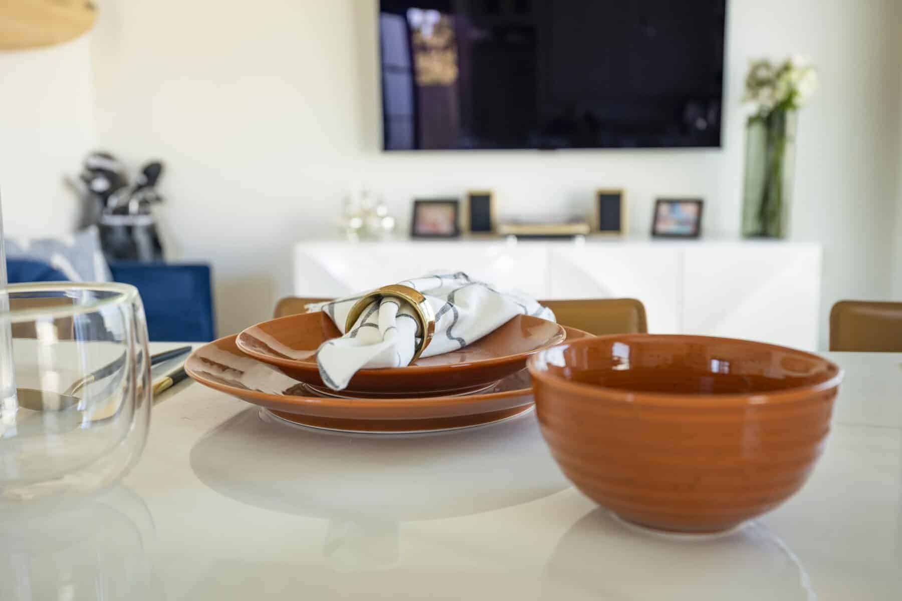Orange dishware and a napkin with a gold ring on a white table, with a modern living area in the background