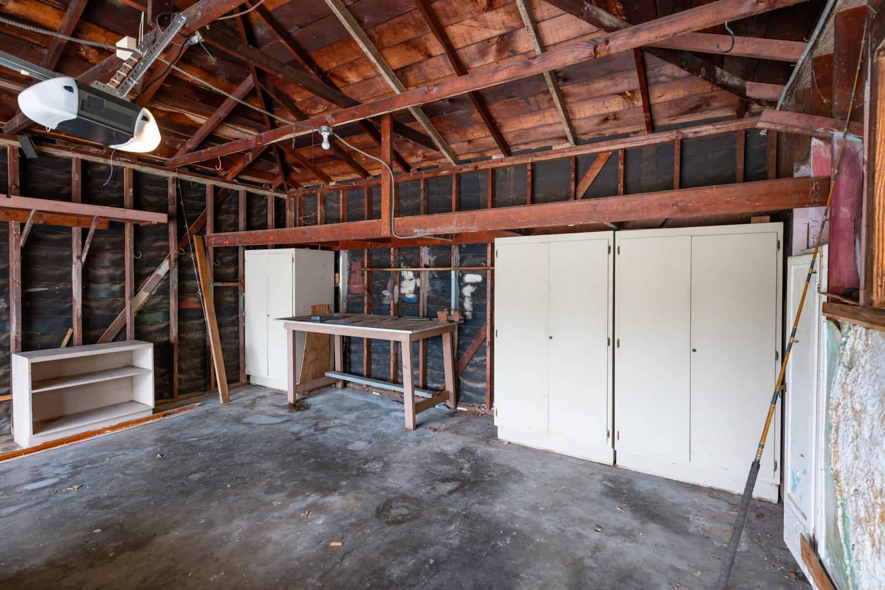 Empty garage with exposed beams, concrete floor, workbench, and built-in white storage cabinets