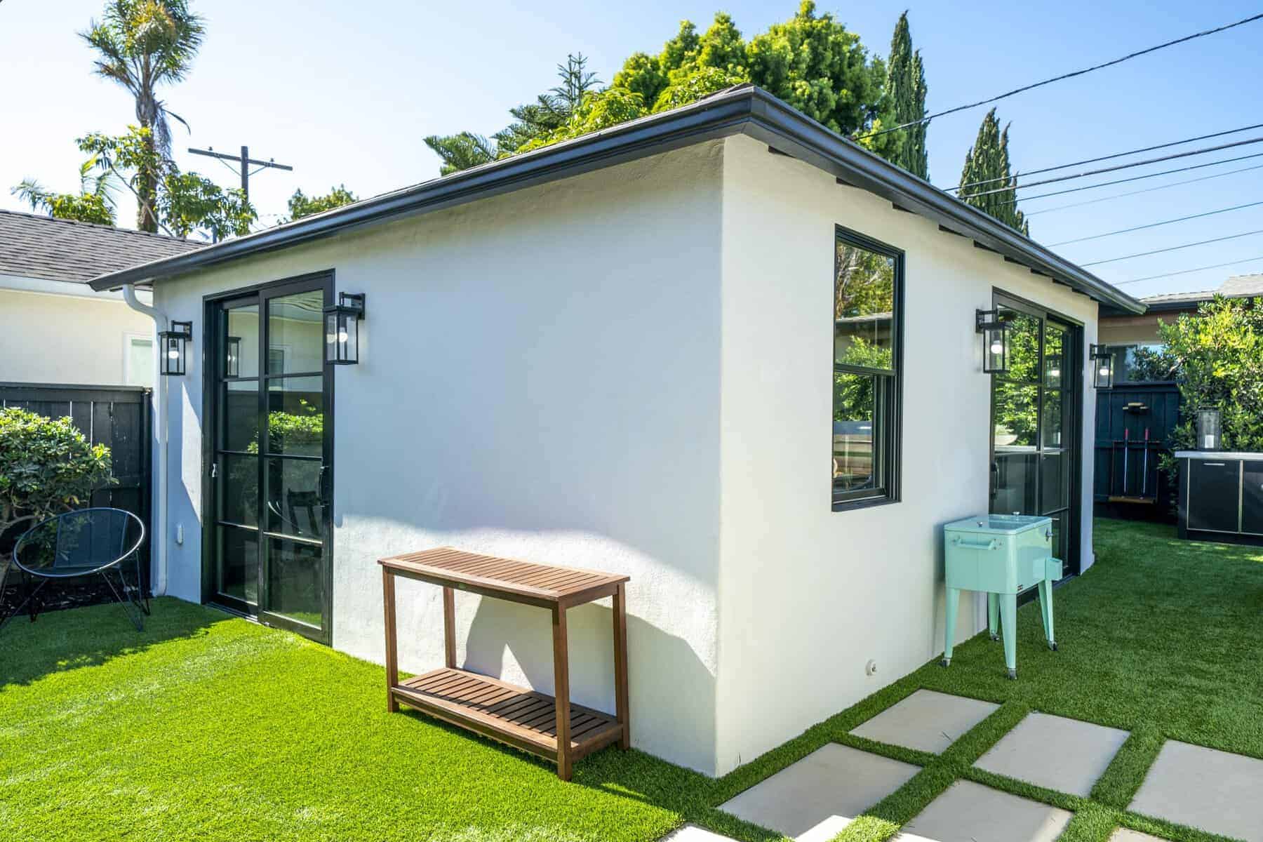 Clean backyard with a white guest house, black-framed glass doors, artificial grass, and minimal outdoor furniture