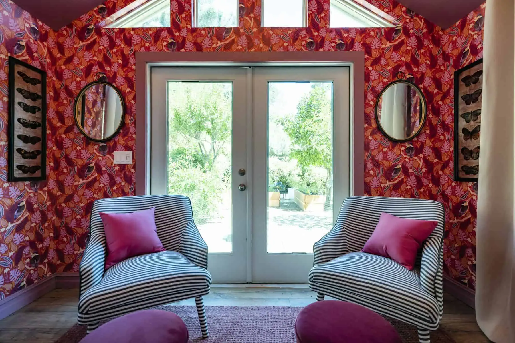 Bold sitting area with tropical wallpaper, striped chairs, pink accents, and French doors opening to a garden view