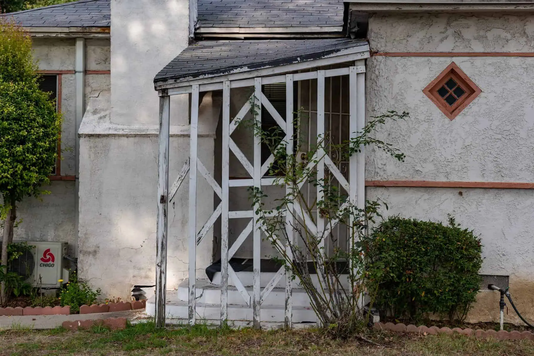 Weathered white stucco house with damaged porch, diamond-shaped window, sloping roof, chimney, shrubs, and trees