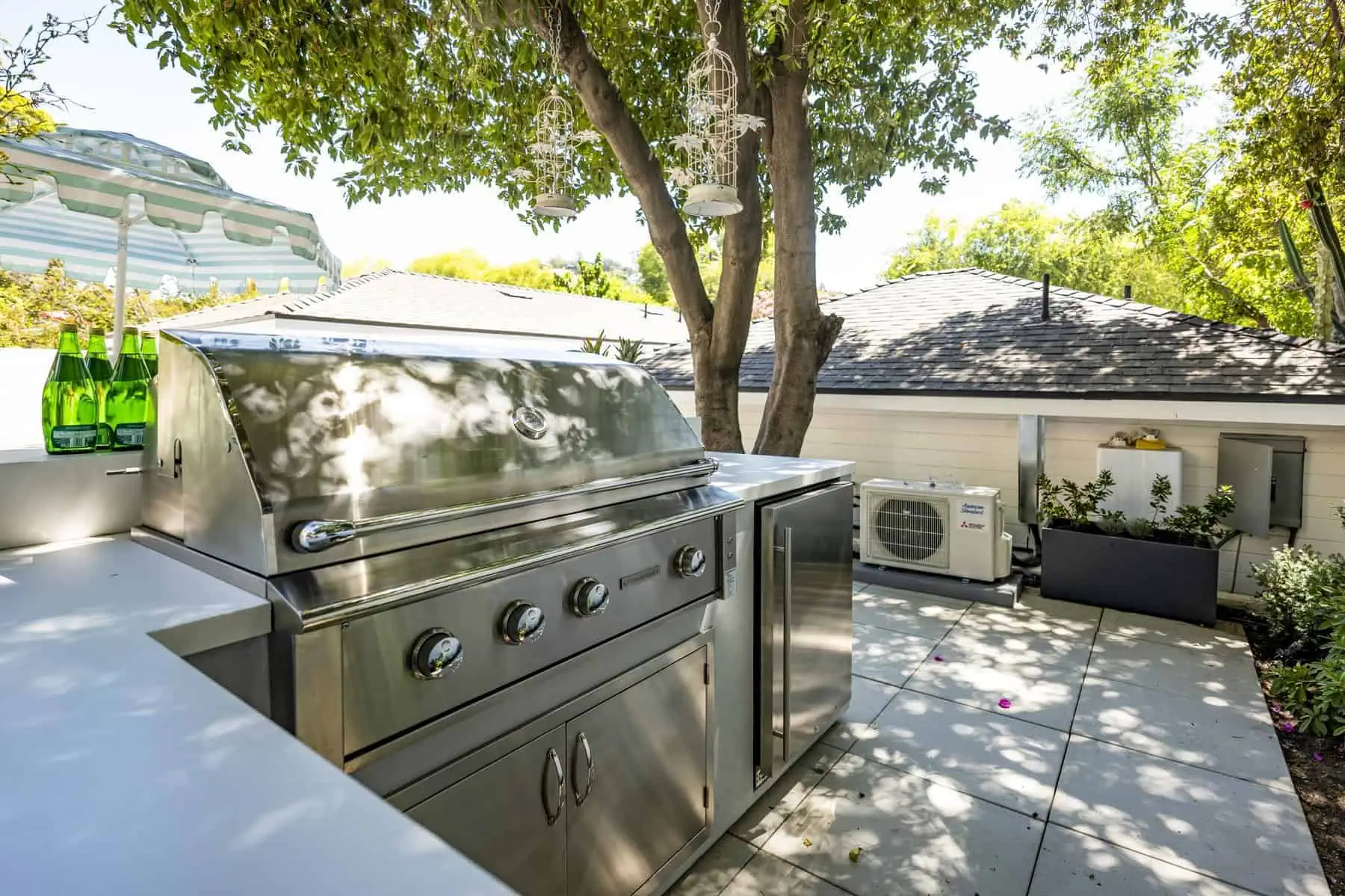 Outdoor kitchen with a stainless steel grill, shaded by a tree, and decorated with lanterns and glass bottles