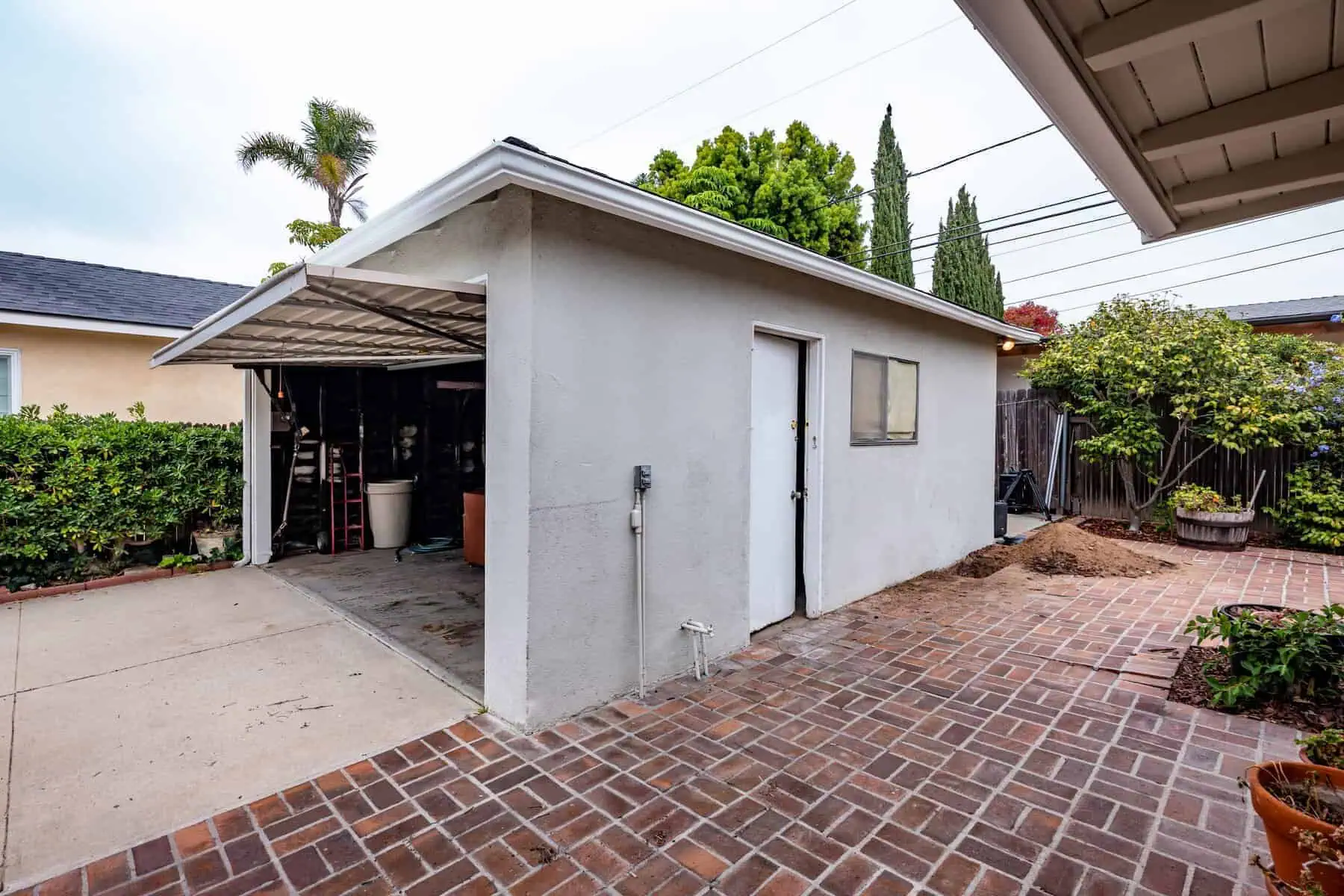 Detached garage with an open door, white exterior, and brick-paved backyard surrounded by greenery