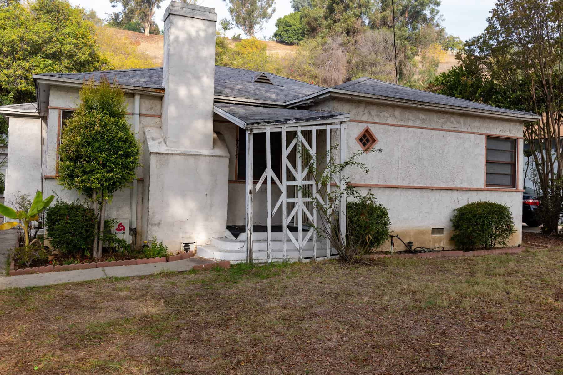 Aging single-story house with stucco exterior, chimney, and a small gated front porch