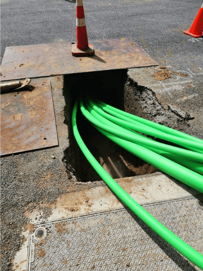 High visibility green utility pipes emerging from underground trench, safety cones on asphalt.