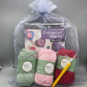 Colourful crochet yarns in pastel green, pink, and red, along with crochet hooks and a crochet pattern book, perfect for crocheted hearts and handmade crafts.