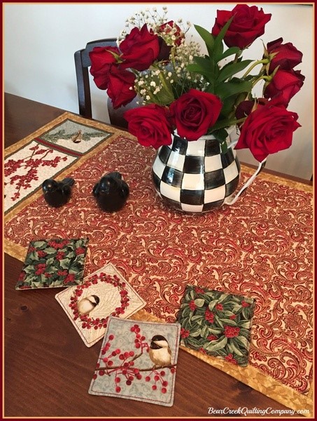 Vase of vibrant red roses with holiday-themed embroidered napkins and decorative ornaments on a rich patterned tablecloth for Christmas décor.