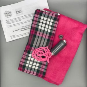 Vibrant pink fleece fabric with matching pink cord and tartan fabric for DIY projects, accompanied by sewing essentials such as thread and a button, perfect for crafted and personalised clothing or accessories.