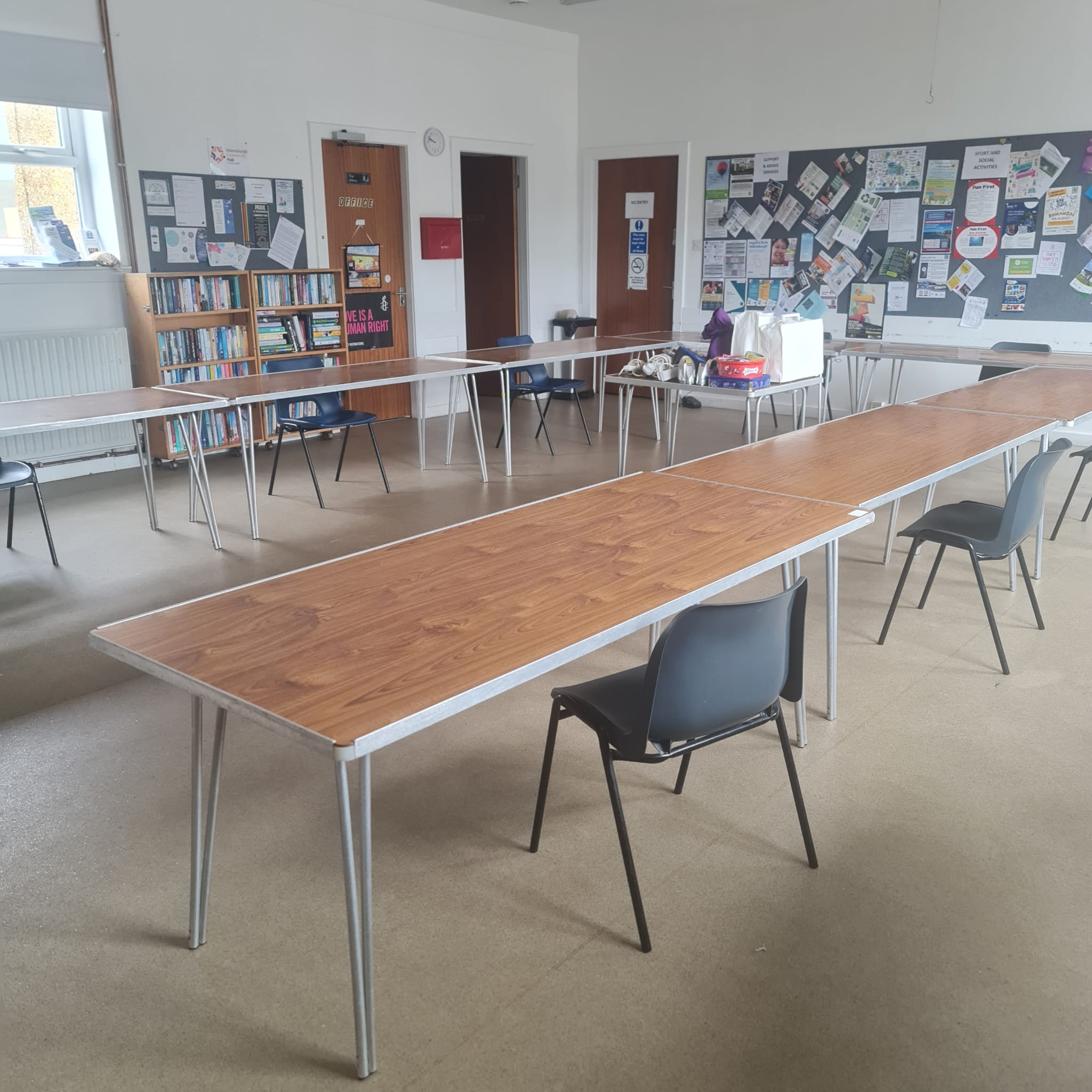 Brightly lit community centre with tables and chairs set up for a meeting or event, featuring a bulletin board, bookshelf, and informational posters in the background.