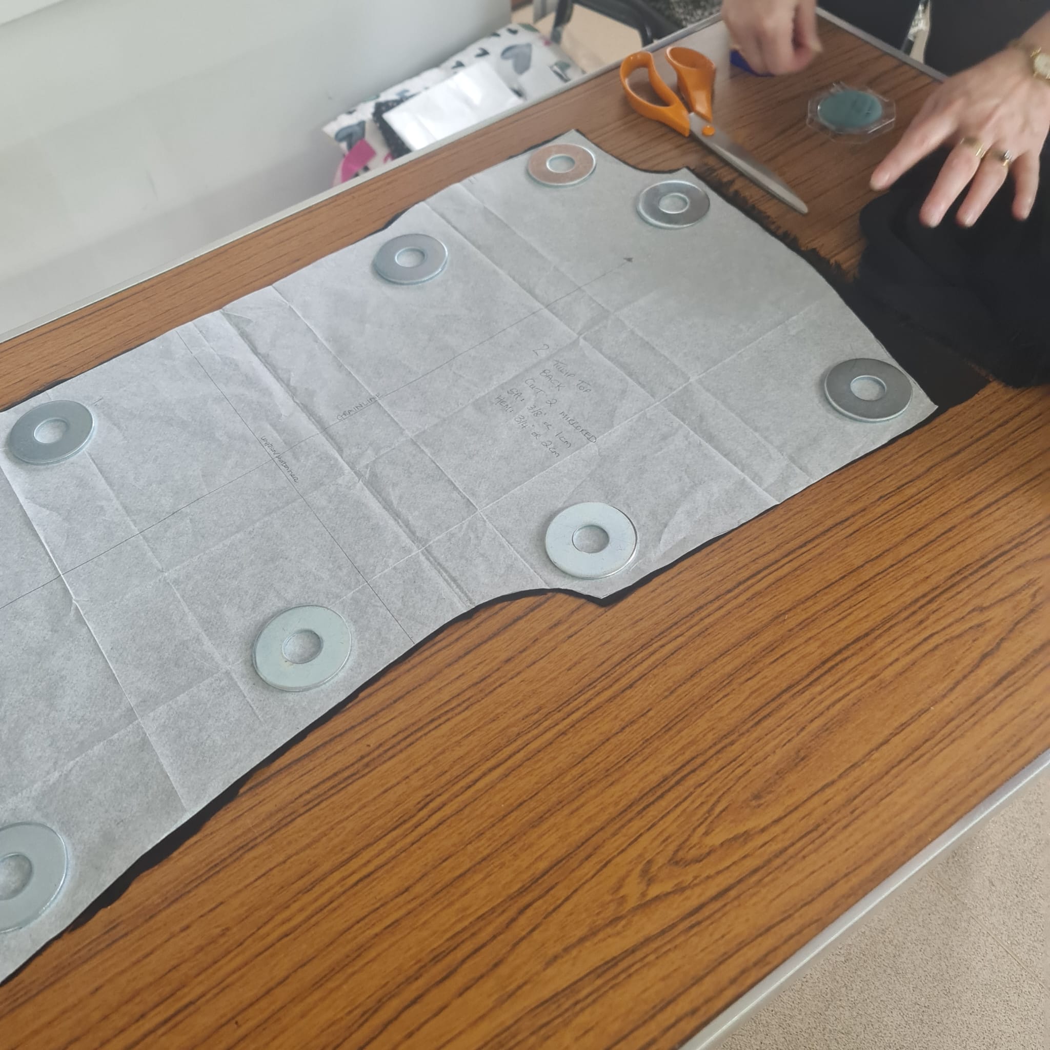 Cutting pattern on parchment paper for garment sewing, with pattern weights to secure fabric, scissors, and measuring tools on a wooden table. Perfect for tailoring, dressmaking, and sewing projects.