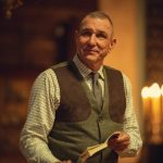 "Middle-aged man in a checkered shirt and vest holding a book, standing indoors with warm lighting, blurred background features candles and wooden decor, creating a cozy, intimate atmosphere."