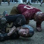 "Two vintage football players wearing old-style helmets and uniforms lie on a muddy field after a tackle, both looking exhausted; blurred players and spectators are seen in the background during a gritty, historical game."