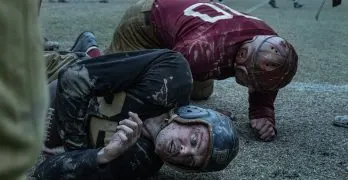 "Two vintage football players wearing old-style helmets and uniforms lie on a muddy field after a tackle, both looking exhausted; blurred players and spectators are seen in the background during a gritty, historical game."