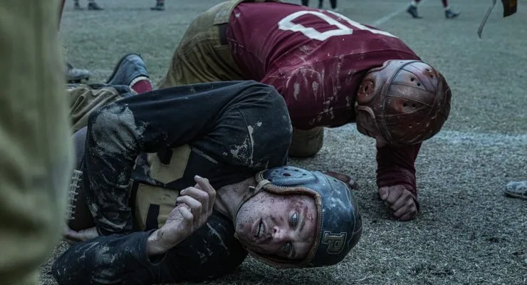 "Two vintage football players wearing old-style helmets and uniforms lie on a muddy field after a tackle, both looking exhausted; blurred players and spectators are seen in the background during a gritty, historical game."