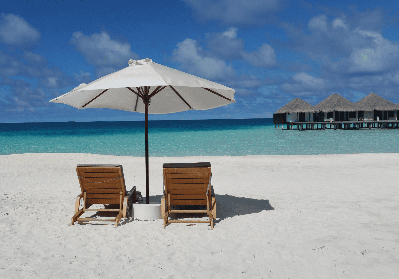 Sunny beach scene with two wooden sun loungers under a large white umbrella on soft white sand, overlooking turquoise waters and overwater bavilions under a partly cloudy sky.