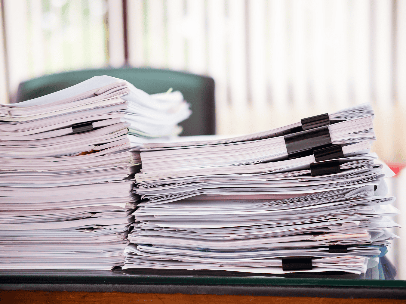 An image of a large stack of disorganized paperwork and files on a desk, with some papers held together by black clips, in an office setting.