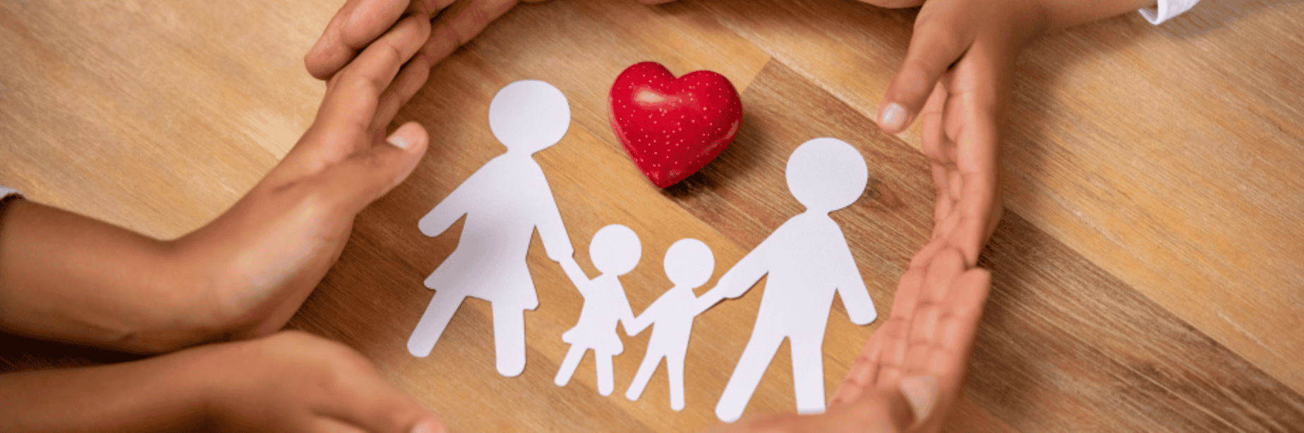 Hand protecting paper family cutouts with a red heart-shaped object on wooden surface.